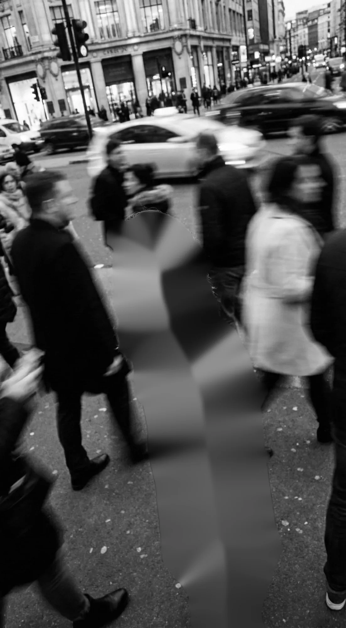 Kait standing still on a busy city crossing, looking back up at the camera with a small smile, in a black coat. The people walking past, the cars and the buildings around her are black and white and blurred by motion. Only she is in colour.