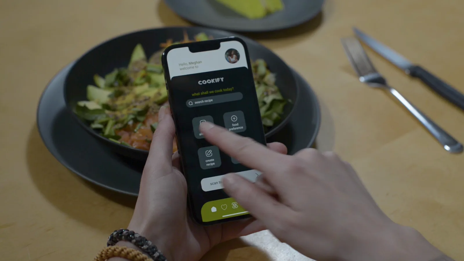 Still from the Cookify film: two hands hold a phone above a dark bowl of salad on a wooden table. The phone shows the Cookify home screen with the wordmark, a search field and four tiles. A knife and fork lie to the right.