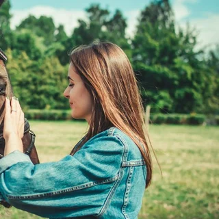 A woman in a denim jacket holding the bridle of a brown horse in a field (photo: Unsplash).