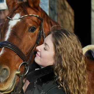 A young woman with long curly hair resting her face against a brown horse by a stable (photo: Unsplash).