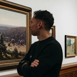 A man with crossed arms looking at a landscape painting in a gallery.