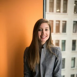 A smiling woman in a grey jacket by a window.