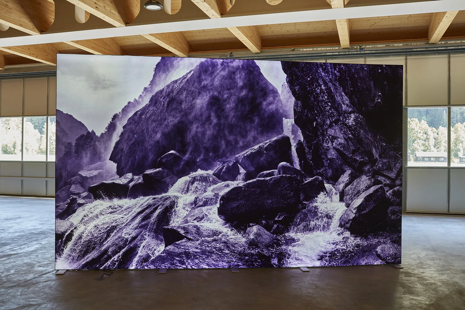 A wall sized photograph of a stream between large rocks, tinted violet, standing as a lit panel on the concrete floor of a hall with a timber roof. Windows and a second panel are behind it.