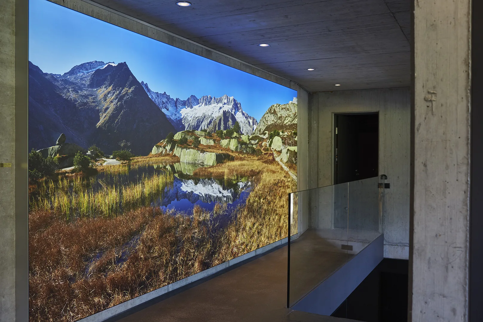 A lit panorama of a mountain lake under a blue sky filling a concrete wall from floor to ceiling beside a glass balustrade.