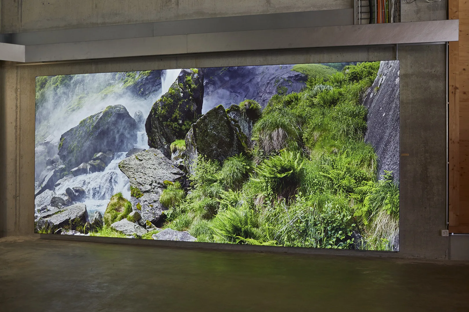 A lit photograph of a waterfall running over rocks between ferns, filling the wall between two concrete pillars.