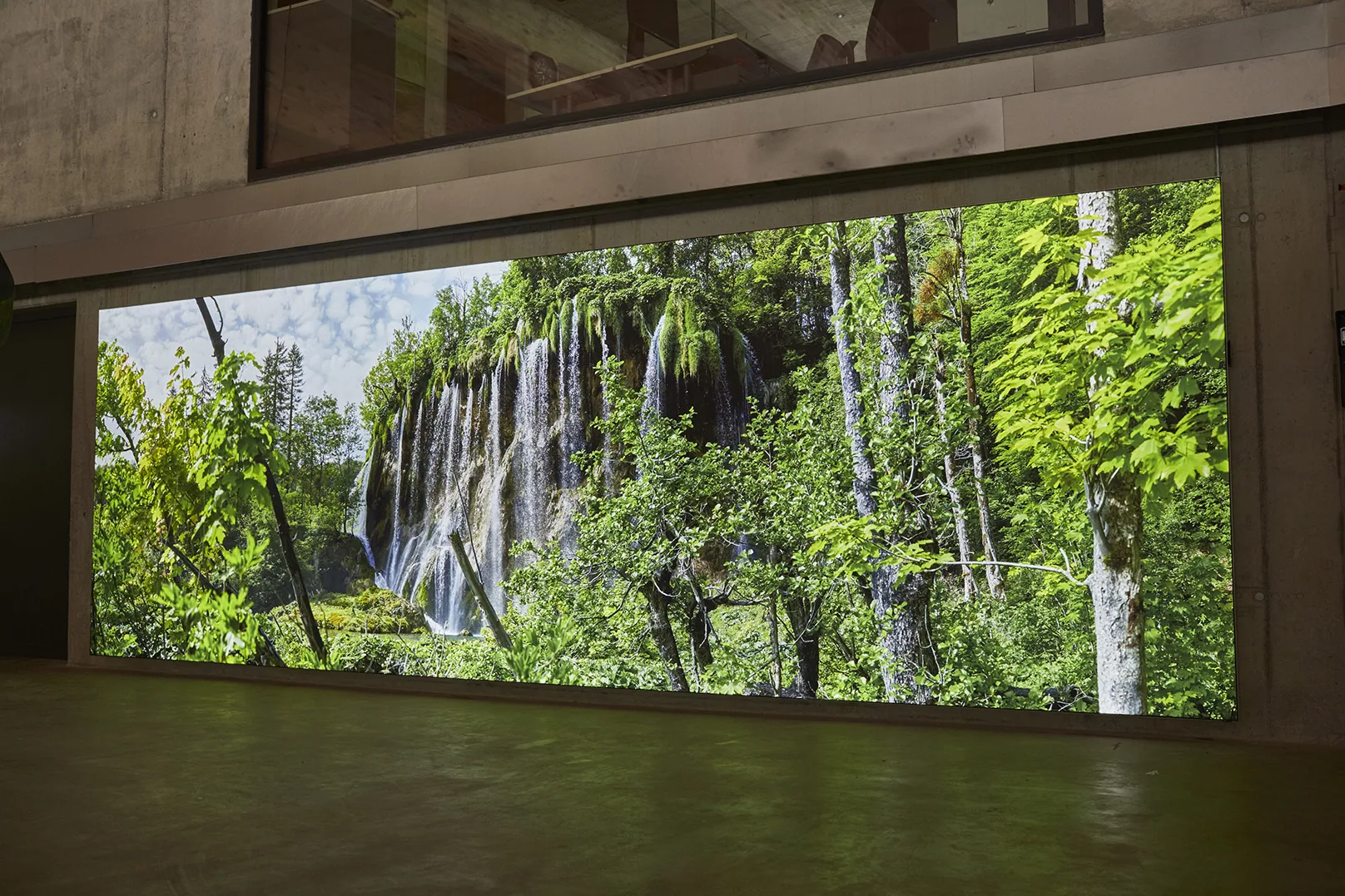 A lit photograph of a wide waterfall in green forest filling the wall under a concrete gallery.