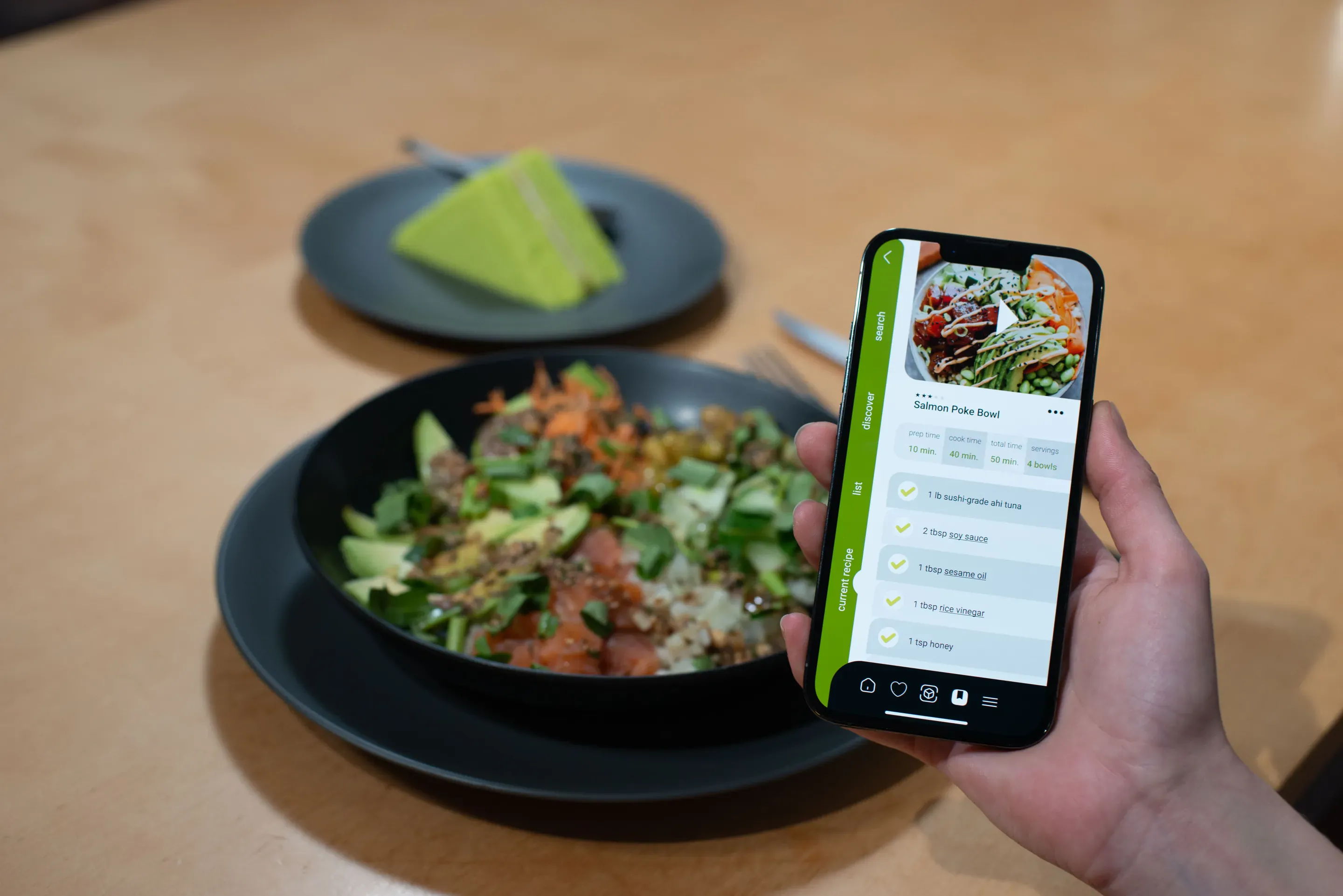 A hand holds a phone above a dark bowl of salad on a wooden table. The phone shows a Cookify recipe screen with a food photo, a checklist of steps and an olive green rail on its left edge. A plate with green slices stands behind.