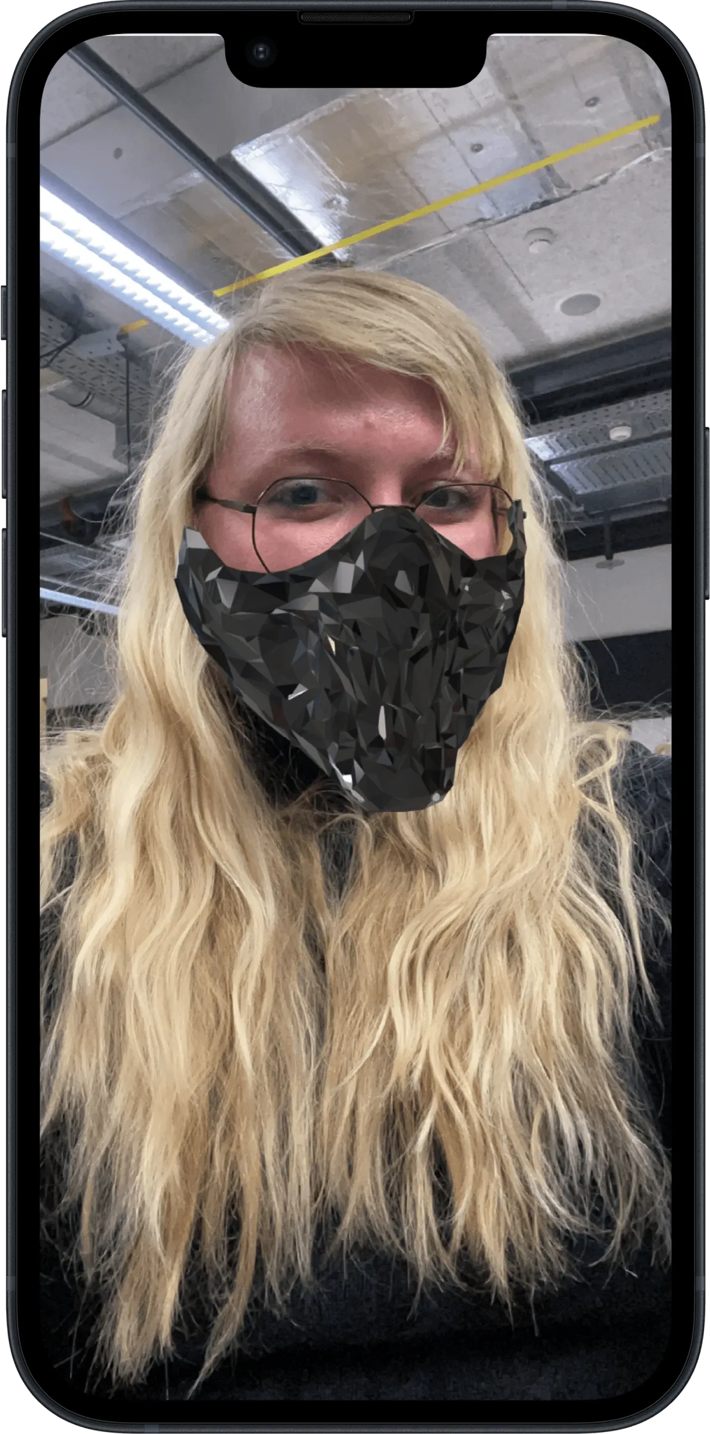 A phone showing the Spark AR prototype: a selfie of a woman with long blonde hair and glasses in a workshop hall, a black faceted mask covers her mouth and nose.