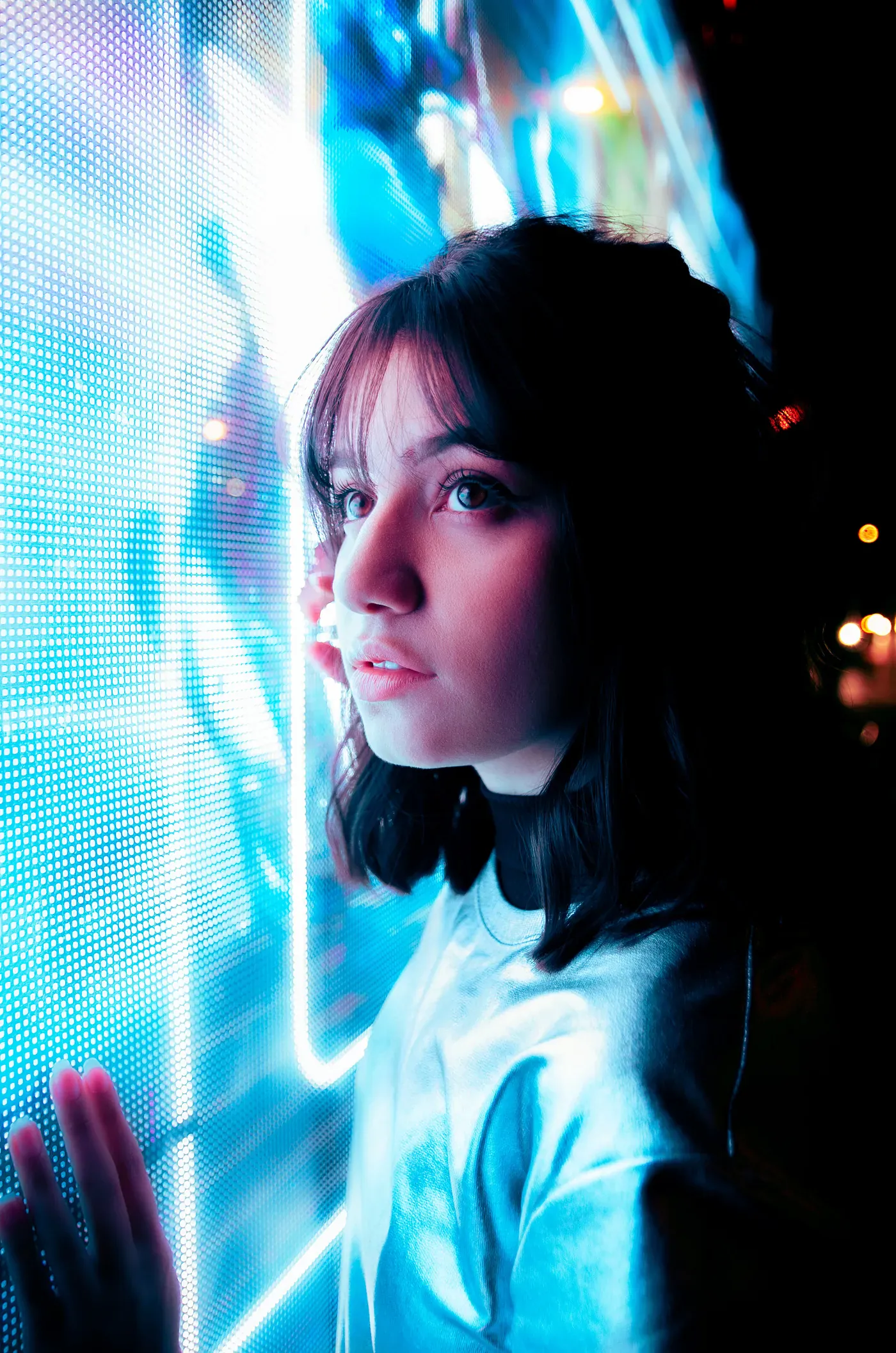 Portrait for the persona: a young woman with dark hair looks to the side, lit by a wall of bright blue screens at night.