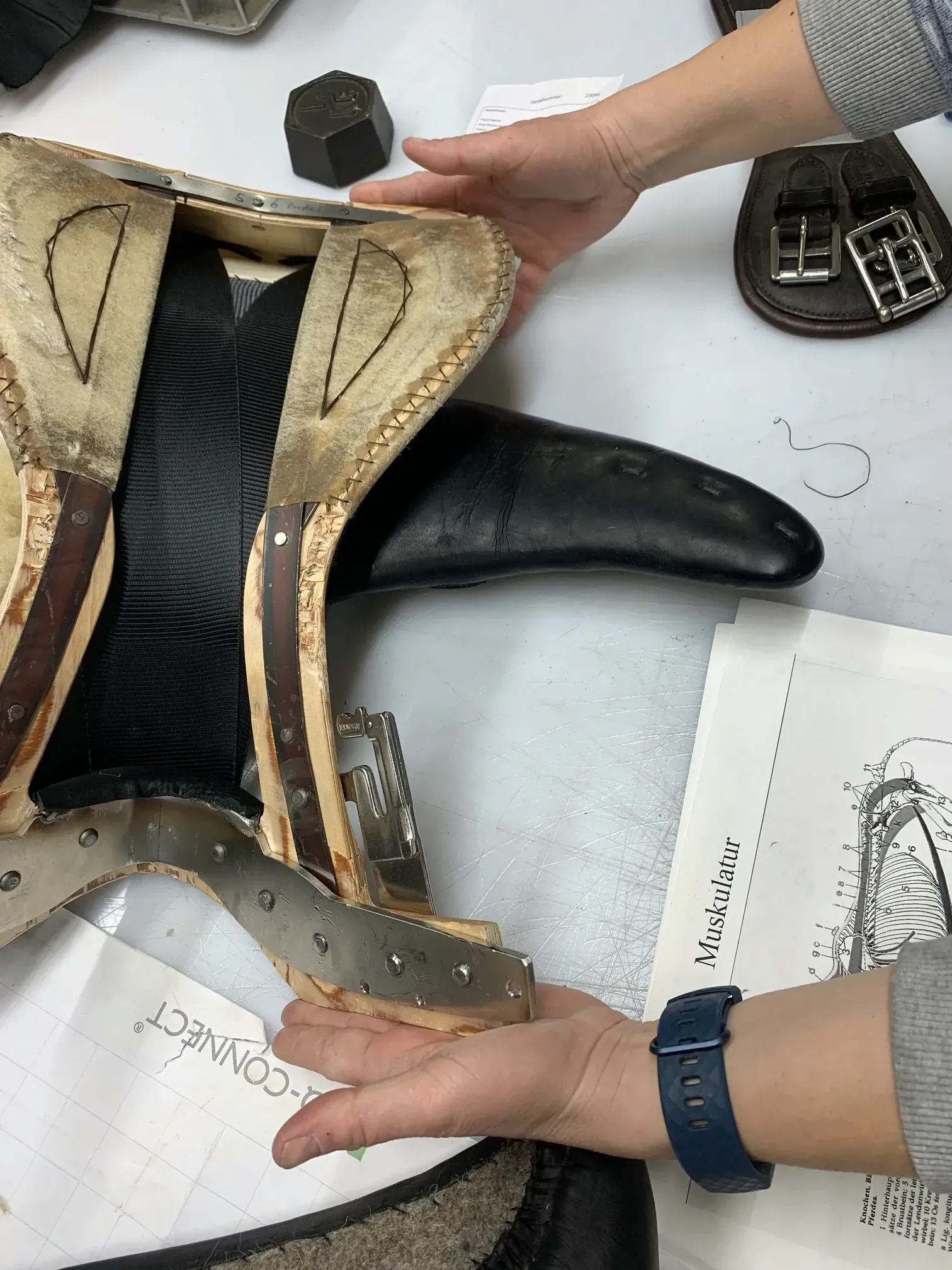 Two people hold an opened saddle on a workbench, showing the tree and padding inside, over technical drawings.