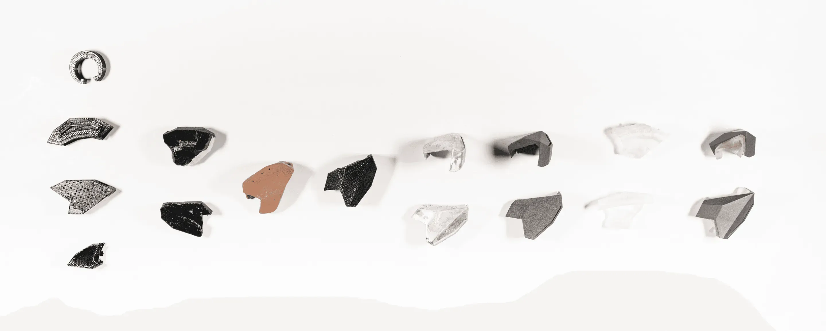 Physical prototypes laid out in rows on white: a ring and about twenty small faceted earbud bodies in black, grey, clear and terracotta material.