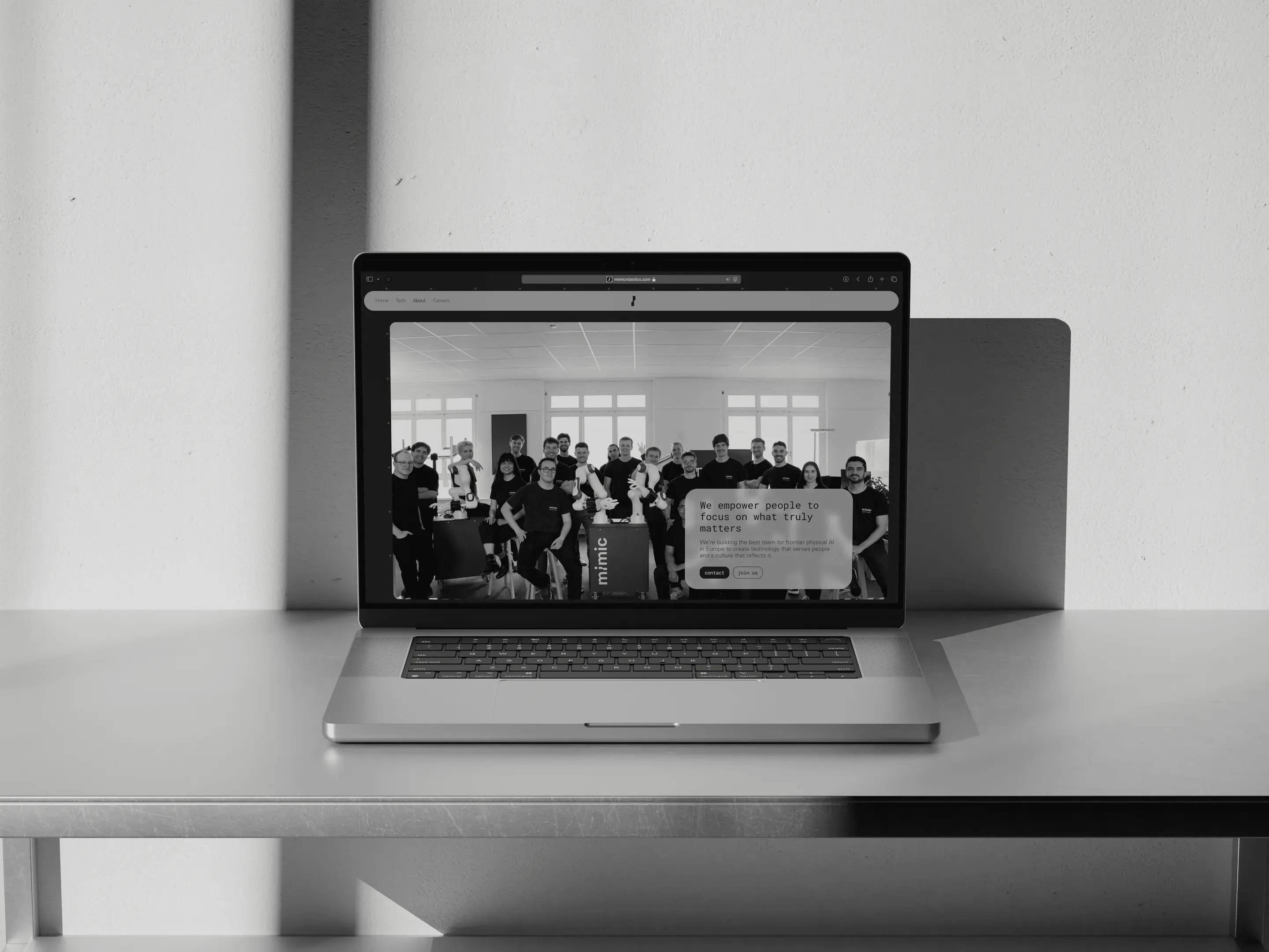 Black and white photo of an open laptop on a table in front of a wall. Its screen shows a website with a group photo of people.