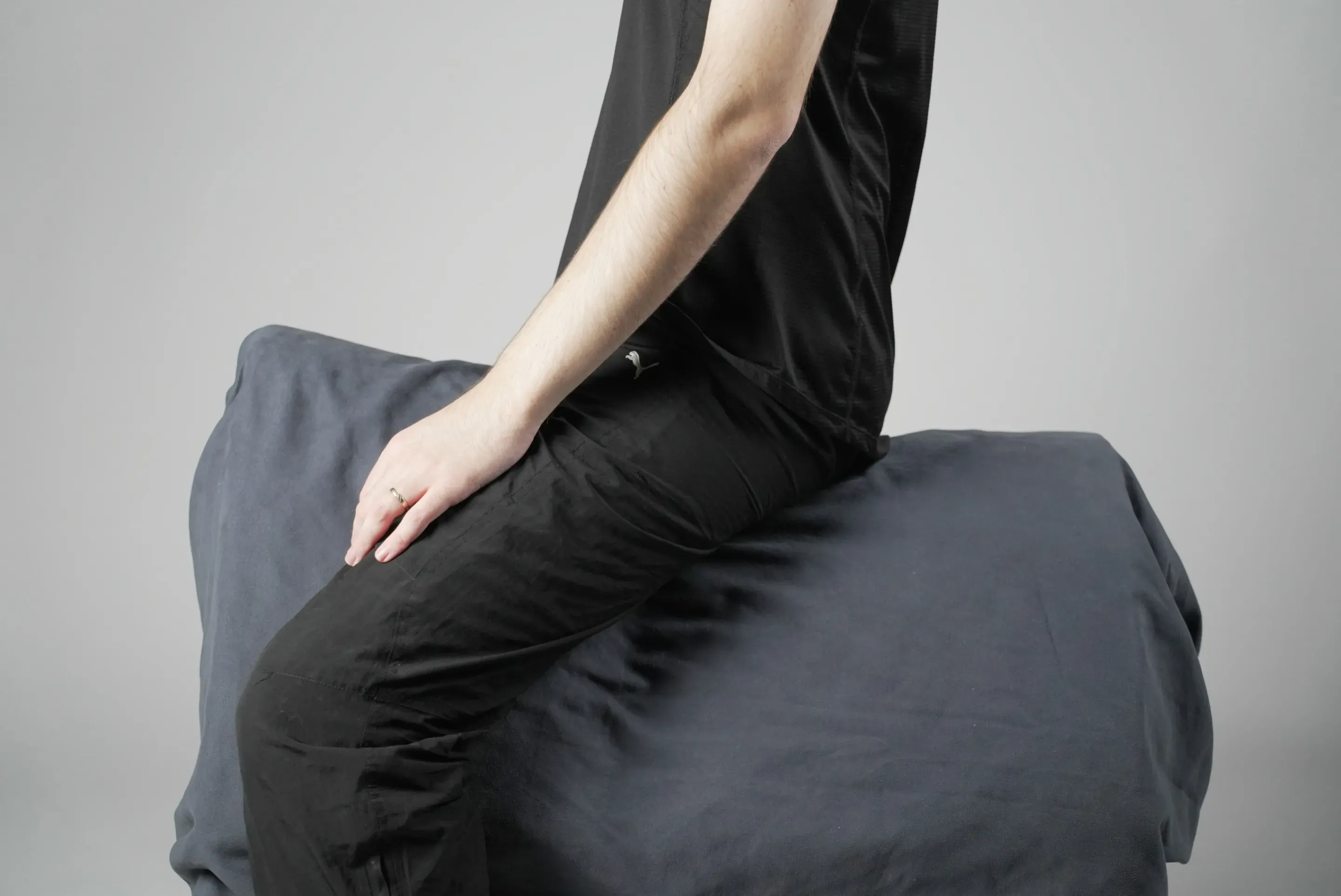 Seat study: a person in black sits astride a padded grey block, seen from the side, leaning slightly back with a hand on the thigh.