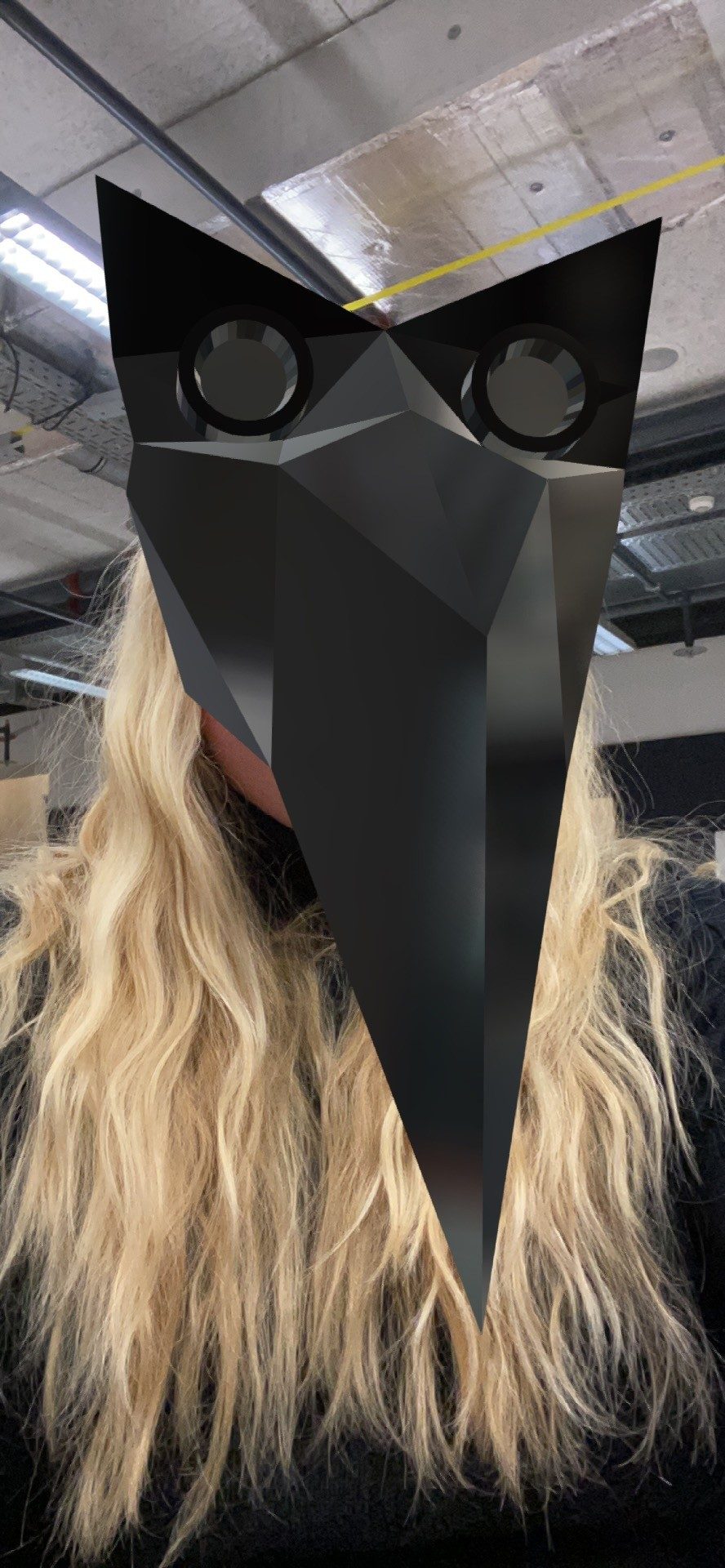 Selfie from the Spark AR prototype in a workshop hall: a woman with long blonde hair, the black mask with a long beak and two round eye pieces covers her face.