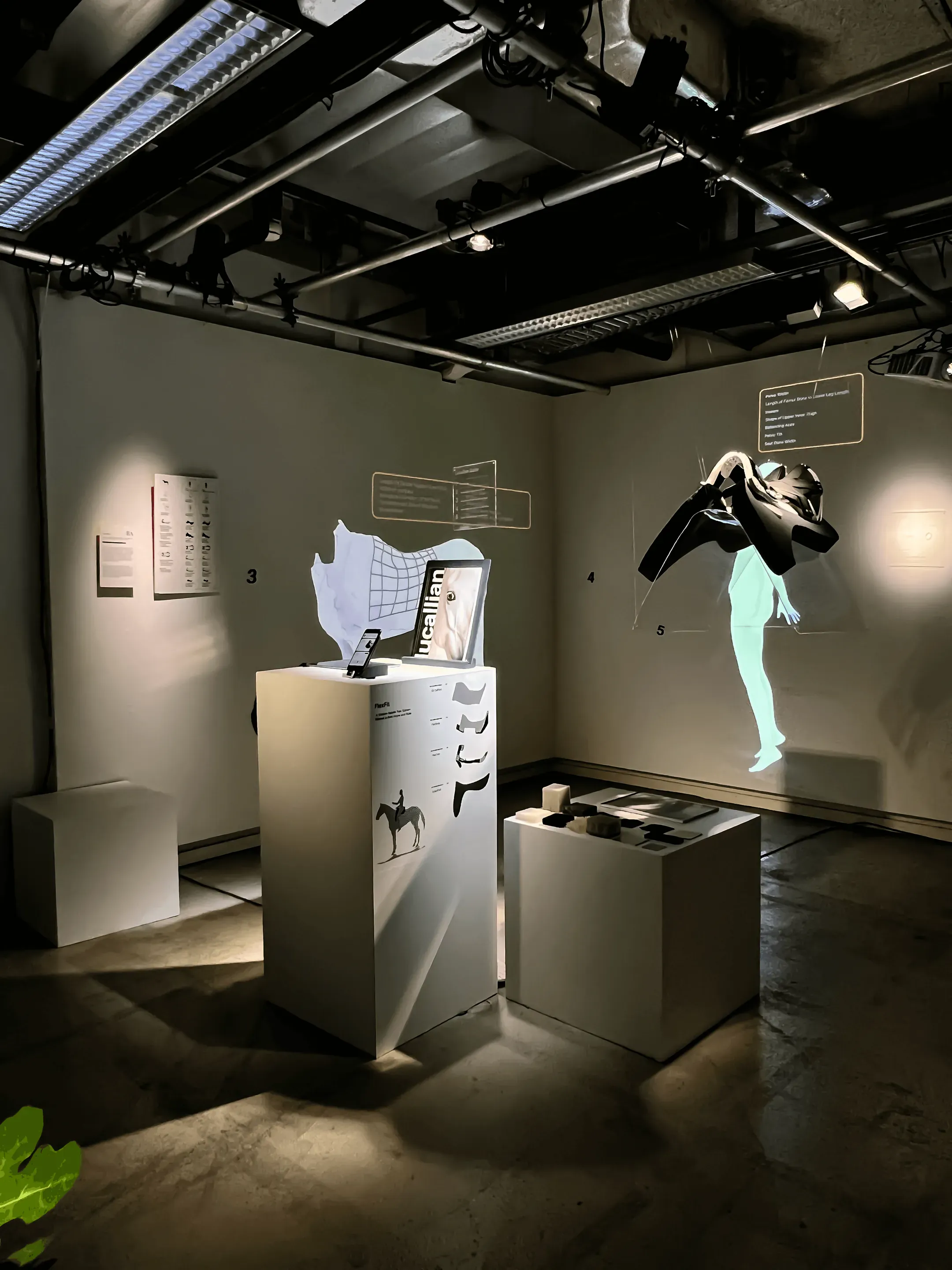 Exhibition stand in a dark room: white plinths with a phone, a Lucallian board and material samples, a saddle tree hanging in the air and a pale figure projected on the wall.